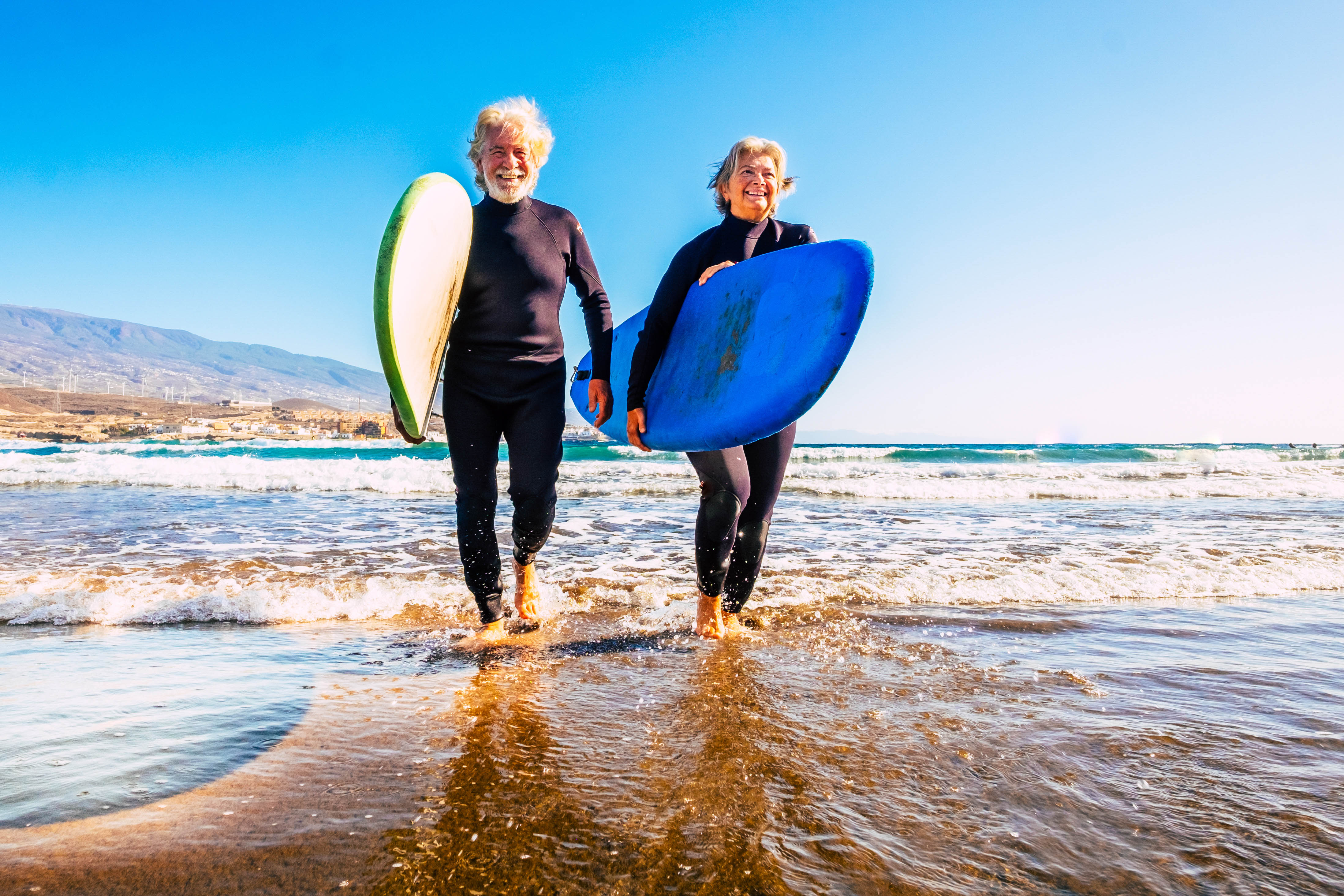 Older couple walking out of the ocean carrying their surfboards and wearing their wet suits, sky is bright blue